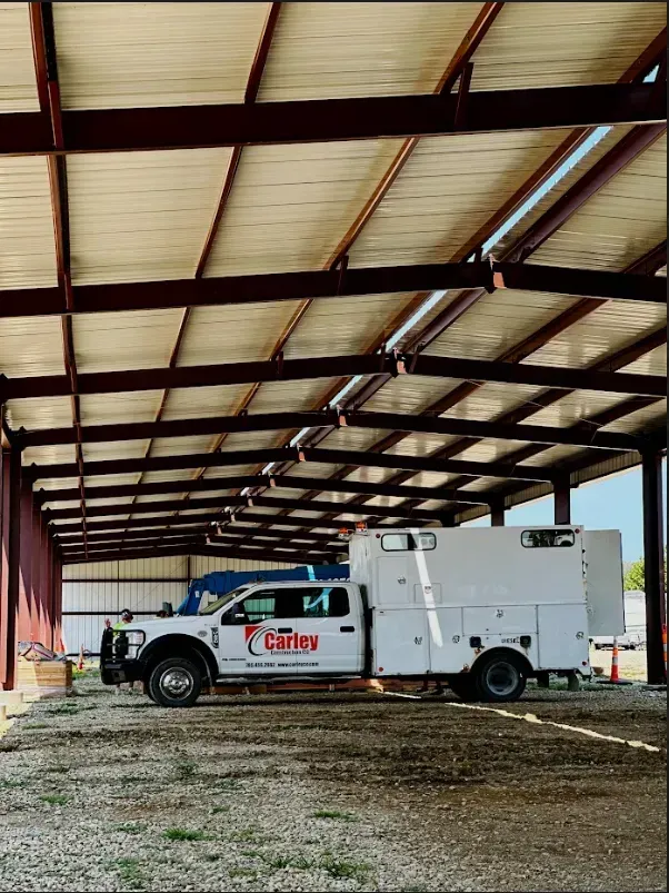 A white work truck parked inside a large, open-air metal warehouse structure with a gravel ground.