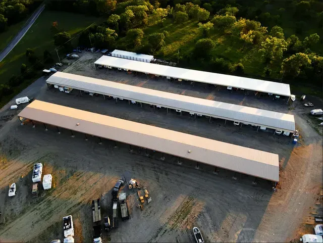 Aerial view of three parallel long, metal-roofed outdoor storage sheds on a gravel lot surrounded by trees.