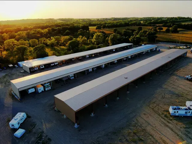 An aerial view of multiple long, metal-roofed storage sheds for RVs and vehicles at sunset near a wooded area.