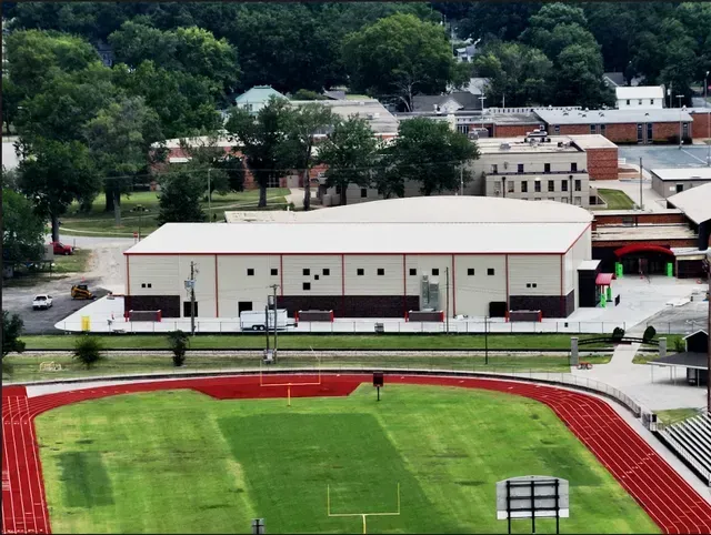 An aerial view of a school athletic facility featuring a bright green football field, red track, and a large gym building.