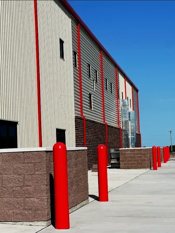 Exterior of a industrial metal building with tan metal walls, red vertical trim, stone base, and a row of red bollards.