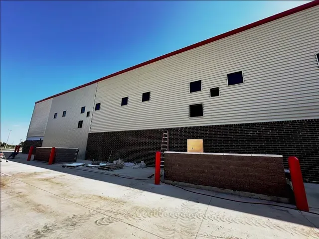 Exterior view of a long building with tan siding and a brick base, featuring scattered square windows under a blue sky.
