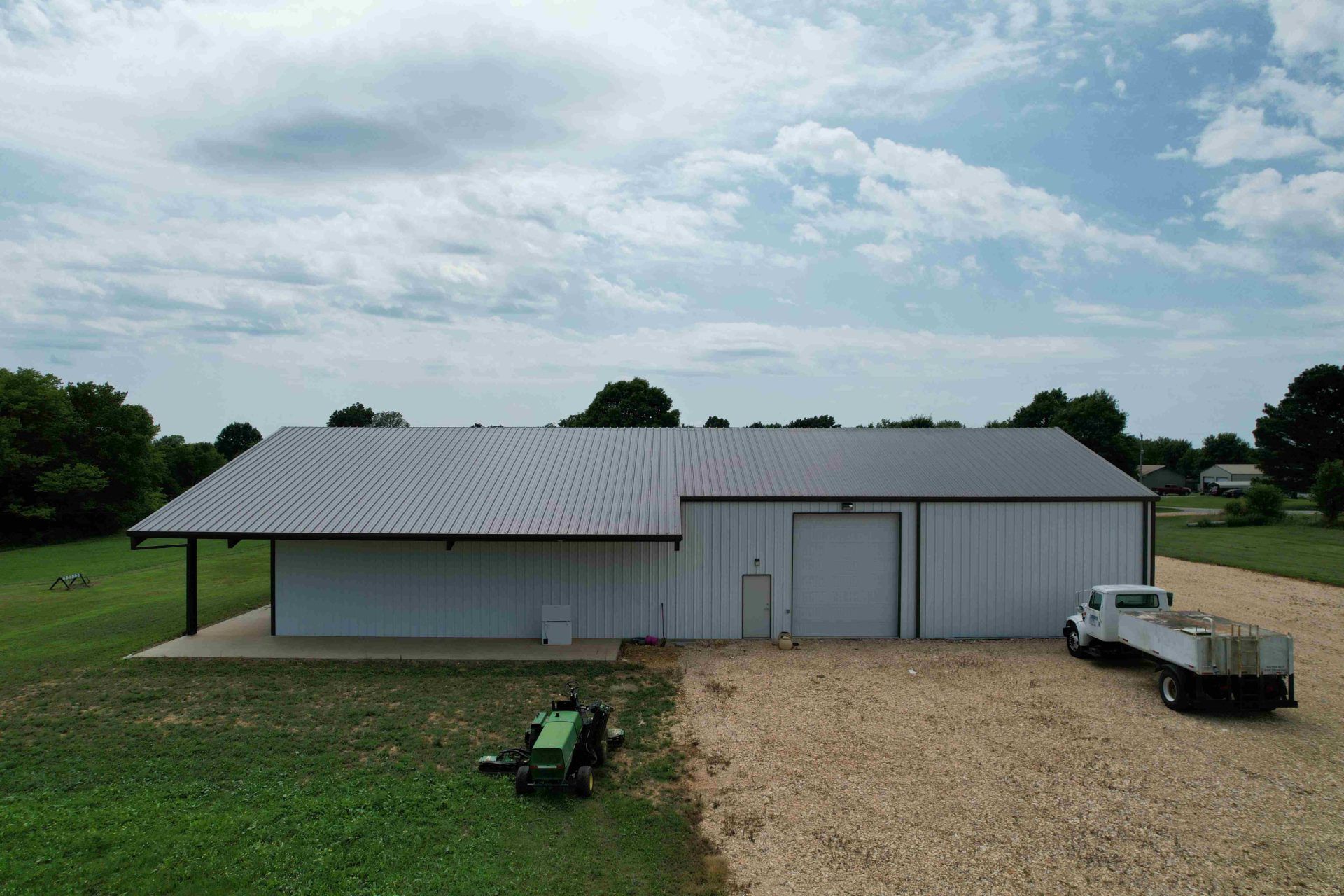 e metal building with sloped roof, loading bay, and truck on gravel driveway with mower on grass