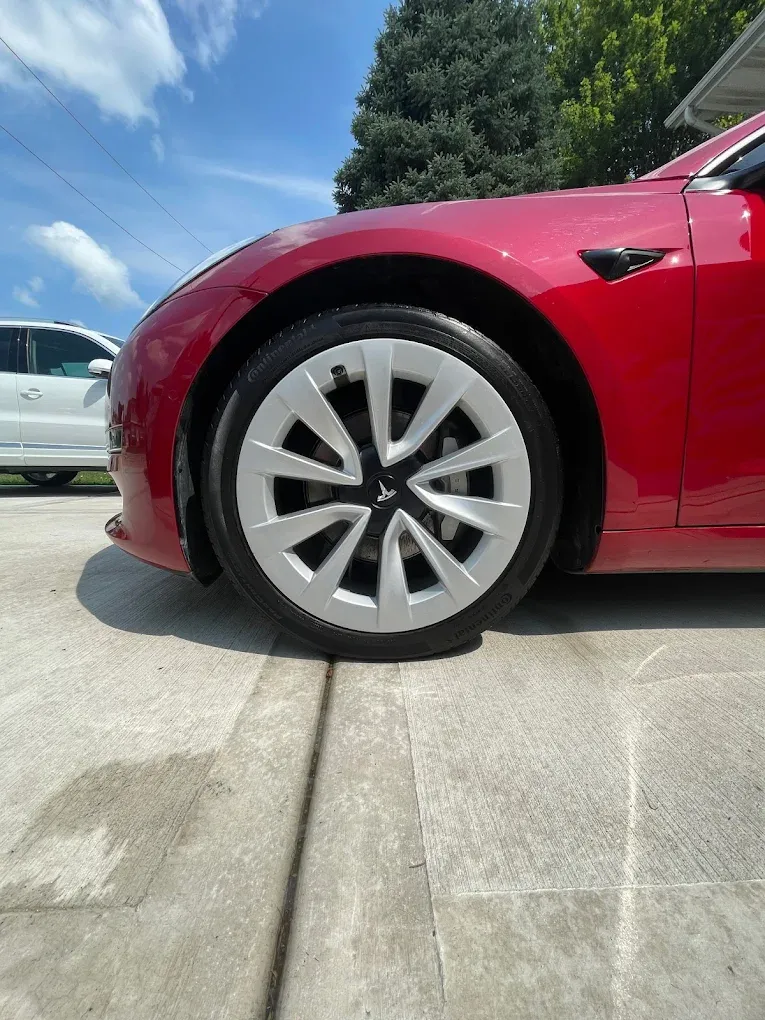 Red car’s front wheel on a driveway, with a white car and trees in the background.
