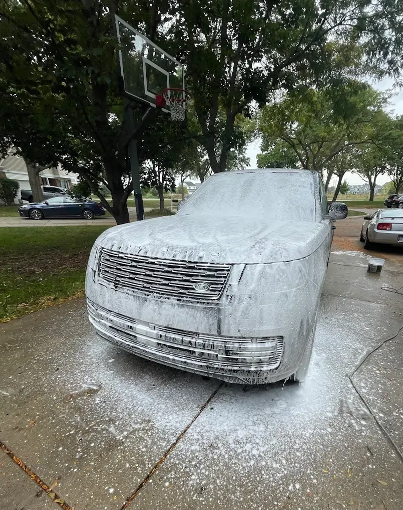 White foam-covered car parked on a wet driveway beneath trees and a basketball hoop.