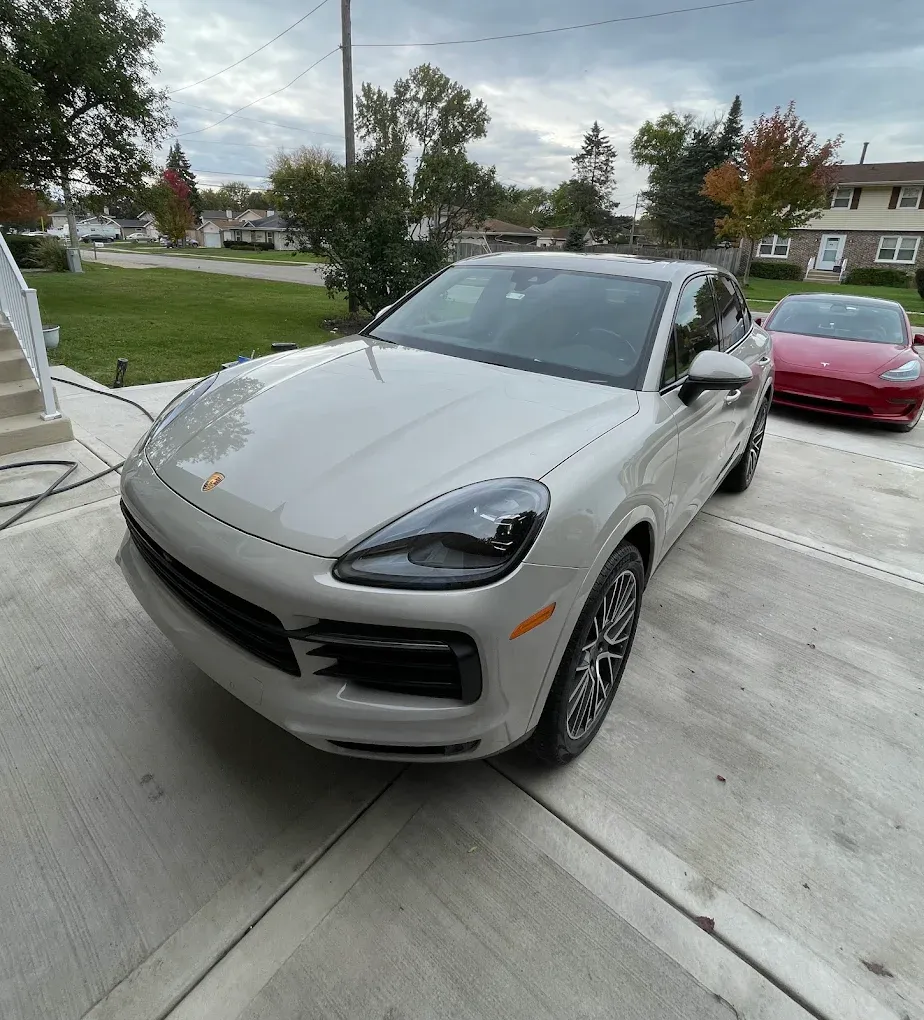 Silver Porsche SUV parked in a driveway, with a red sports car behind it and cloudy skies overhead.