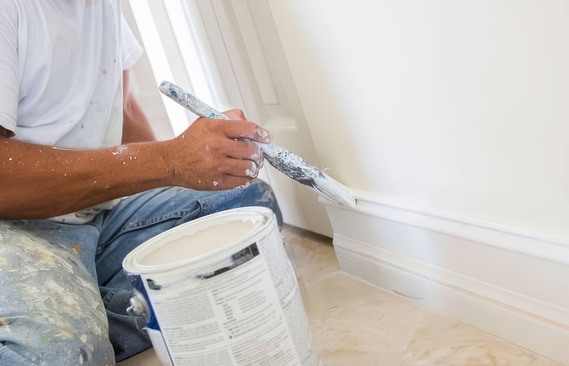 Person painting a white wall trim with a paintbrush beside a paint bucket Person painting a white wall trim with a paintbrush beside a paint bucket