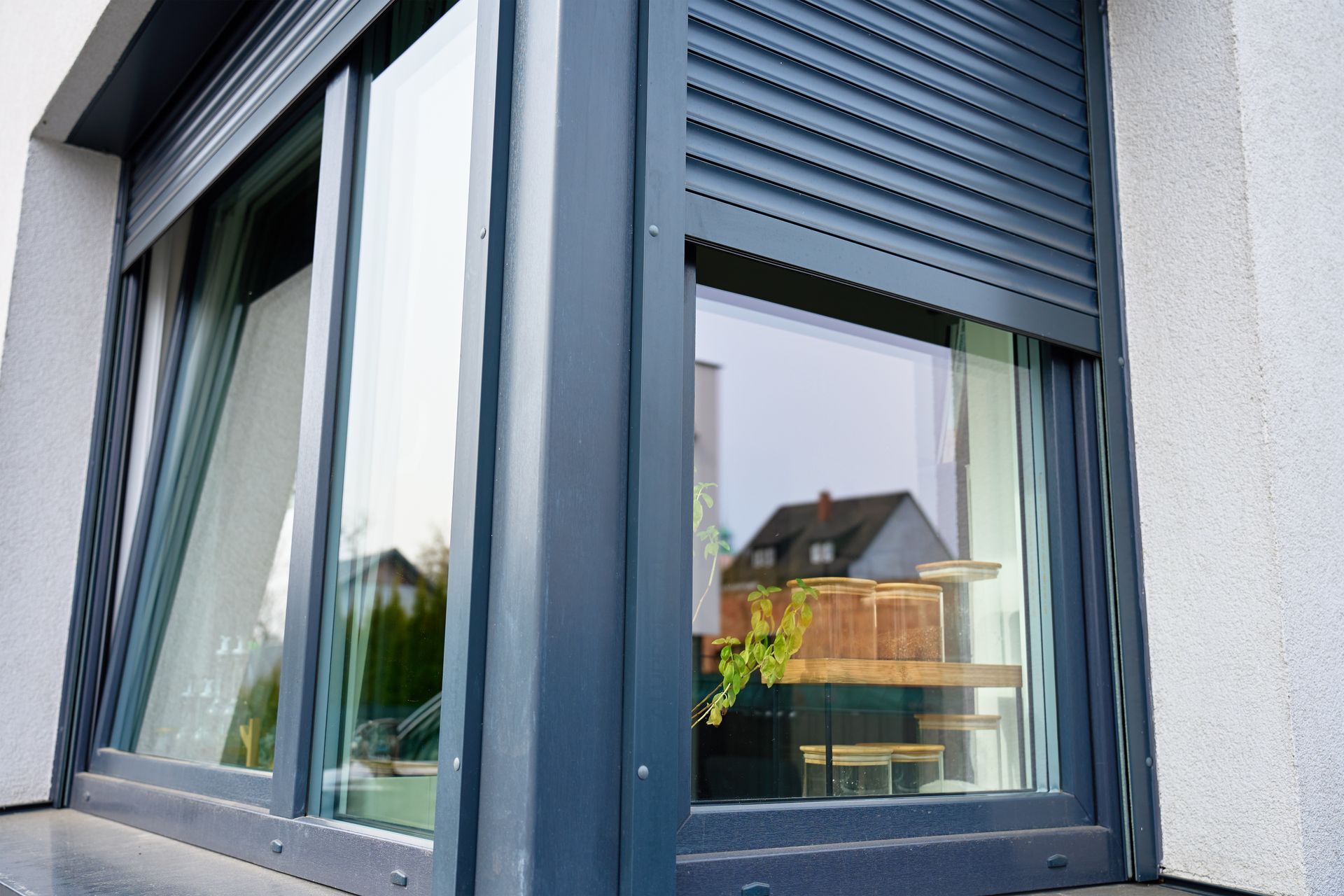 Corner of a modern house with a blue-framed window and partially closed gray shutters reflecting a building and plants Corner of a modern house with a blue-framed window and partially closed gray shutters reflecting a building and plants