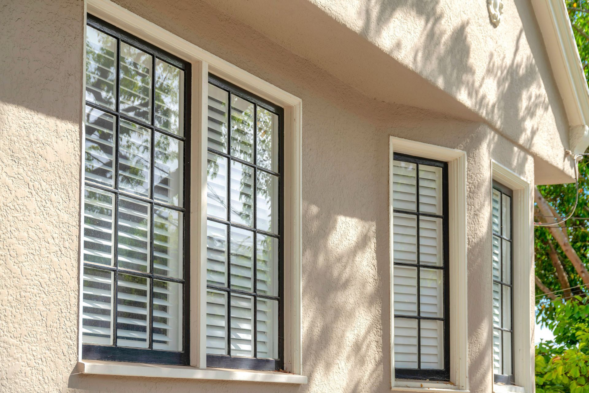 Sunlit beige house exterior with three windows and white shutters, with tree shadows on the wall Sunlit beige house exterior with three windows and white shutters, with tree shadows on the wall