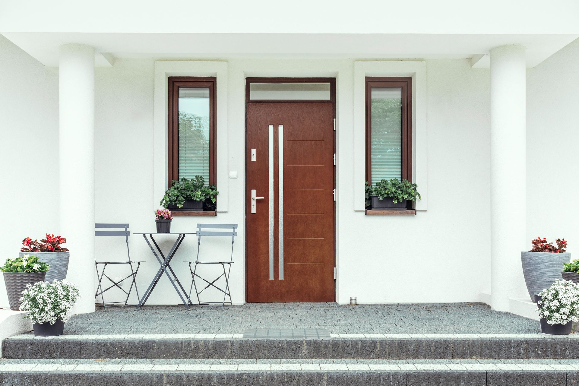 Modern white house entrance with brown front door, narrow windows, and potted plants on either side.