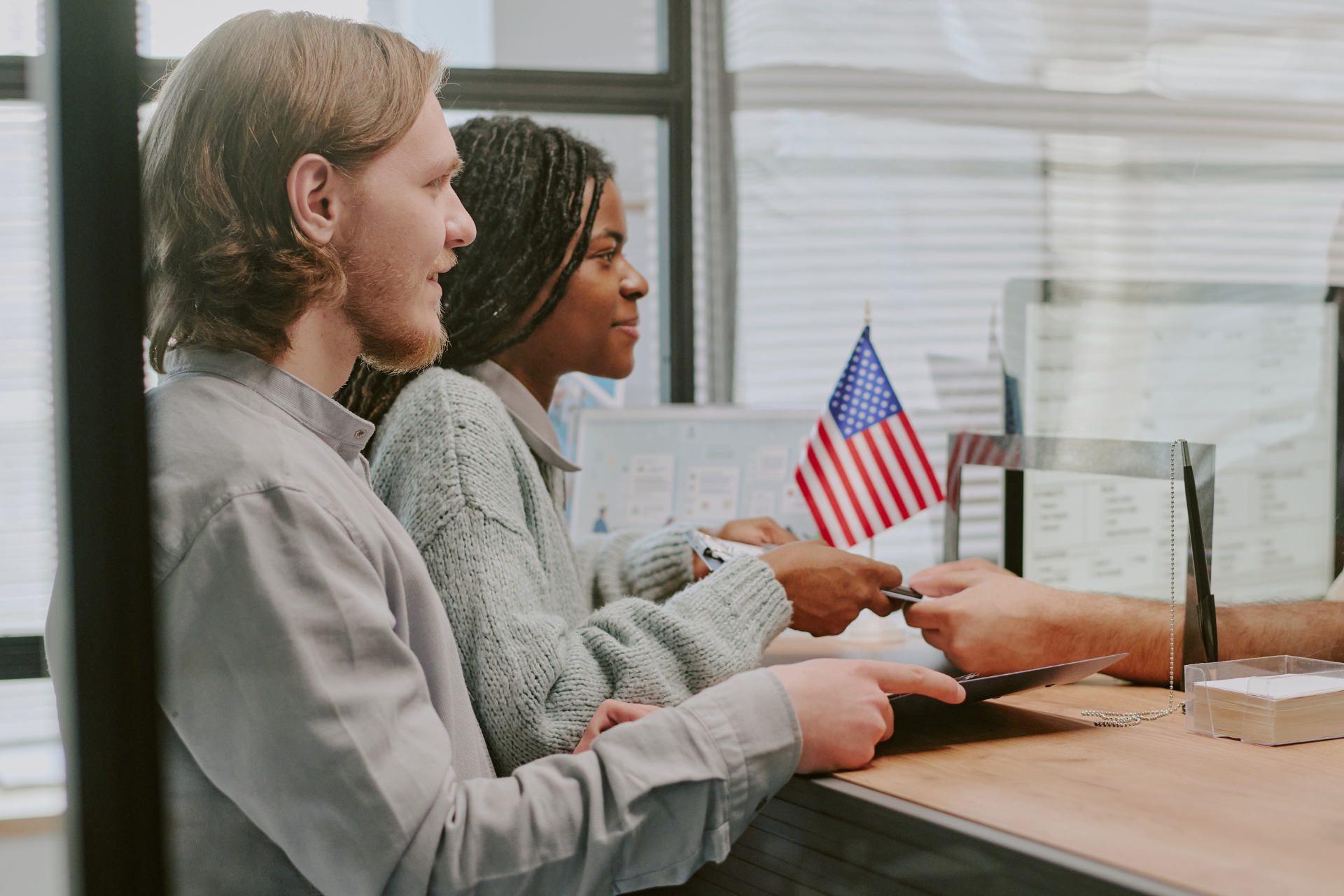 Two people seated at a counter in an office, exchanging papers beside a small American flag.