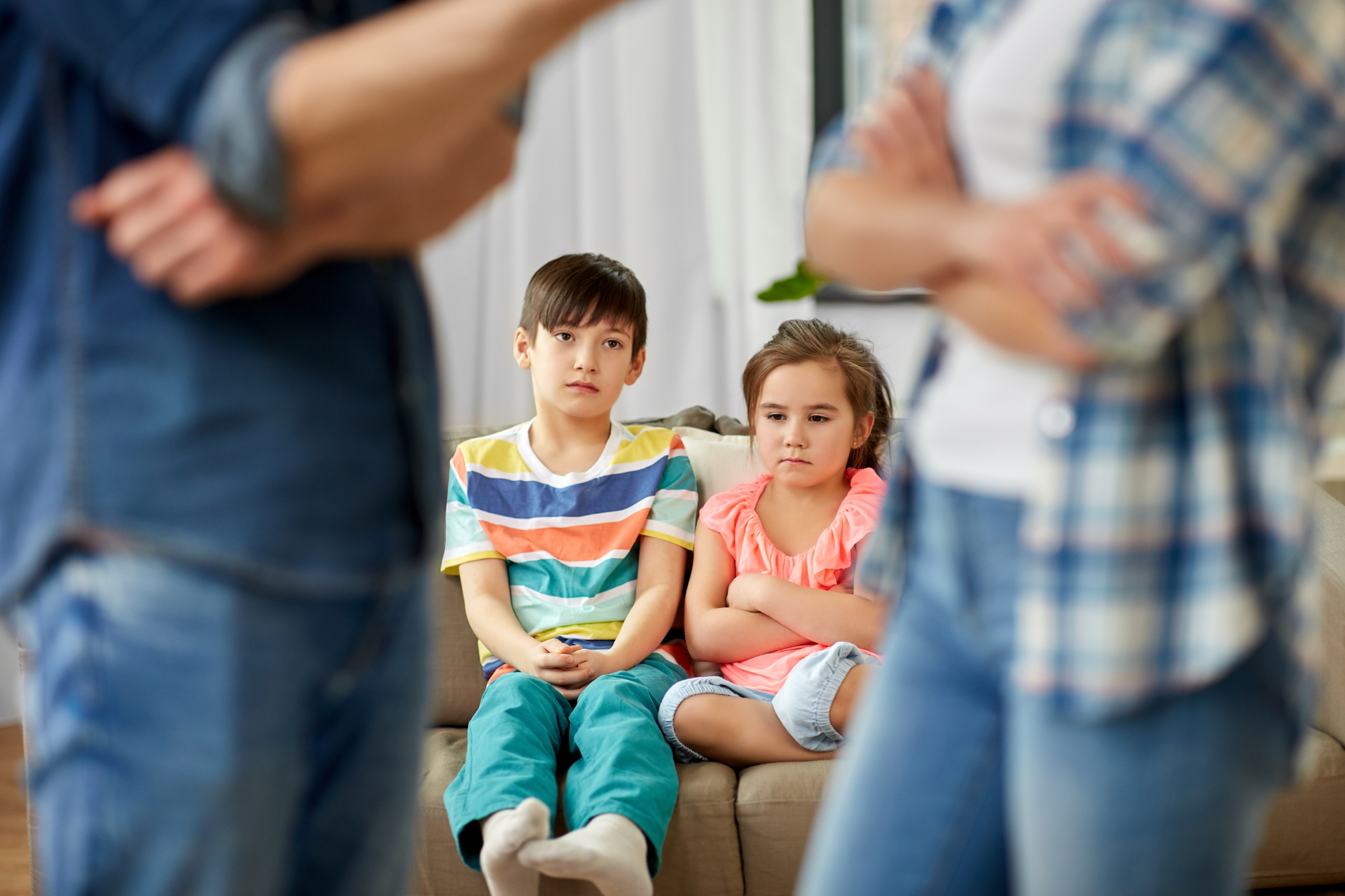 Two children sitting together, blurred adults standing nearby in a bright indoor setting