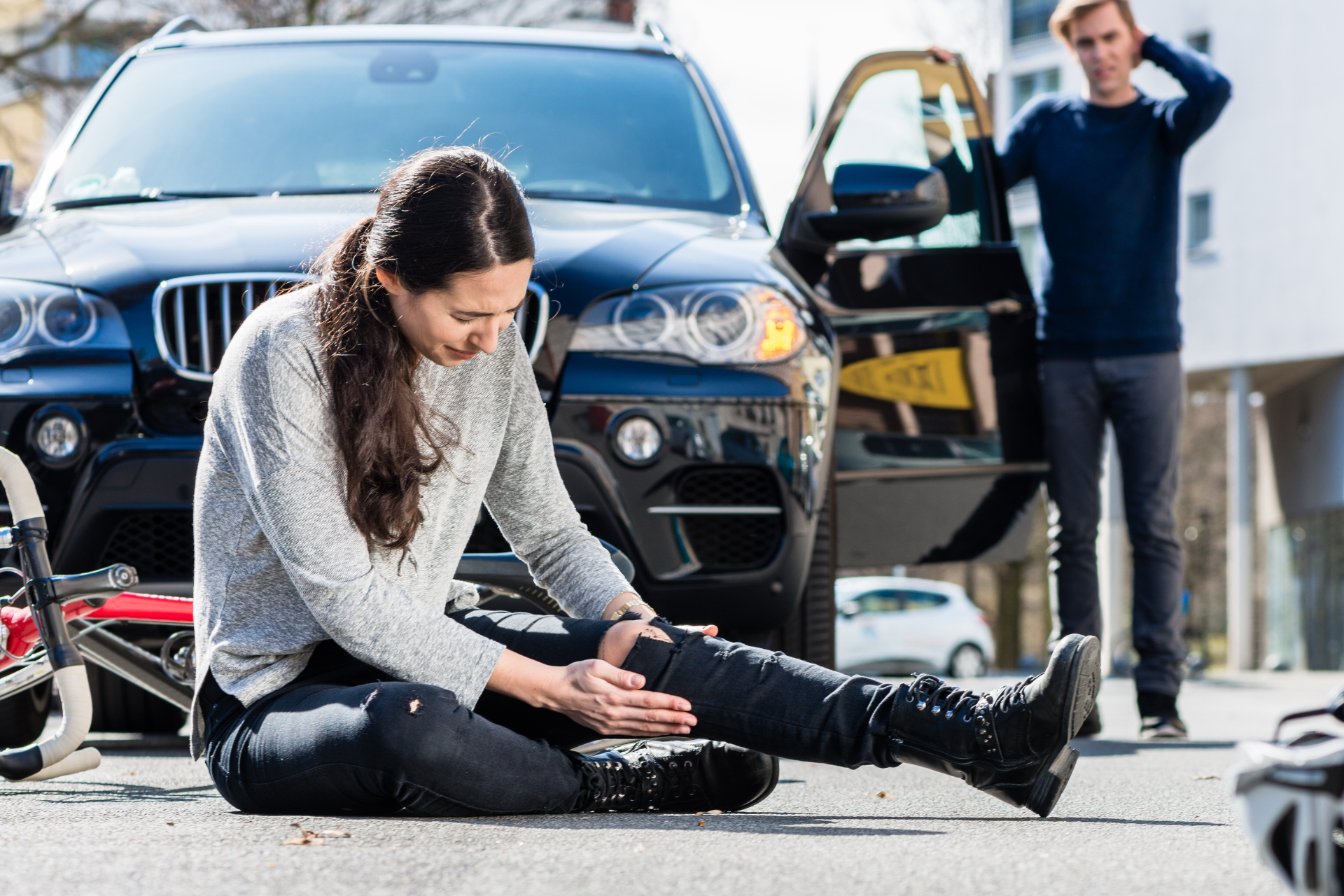 Person sitting on pavement with injured leg in front of a black car; another person stands nearby.