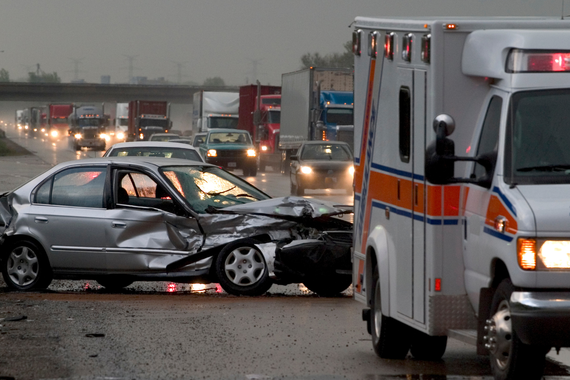 Damaged car after collision beside an ambulance on a wet road, with emergency vehicles in the background