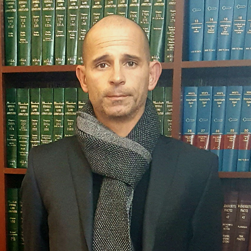 Man in dark suit and gray scarf standing before a bookshelf of green and blue law books.