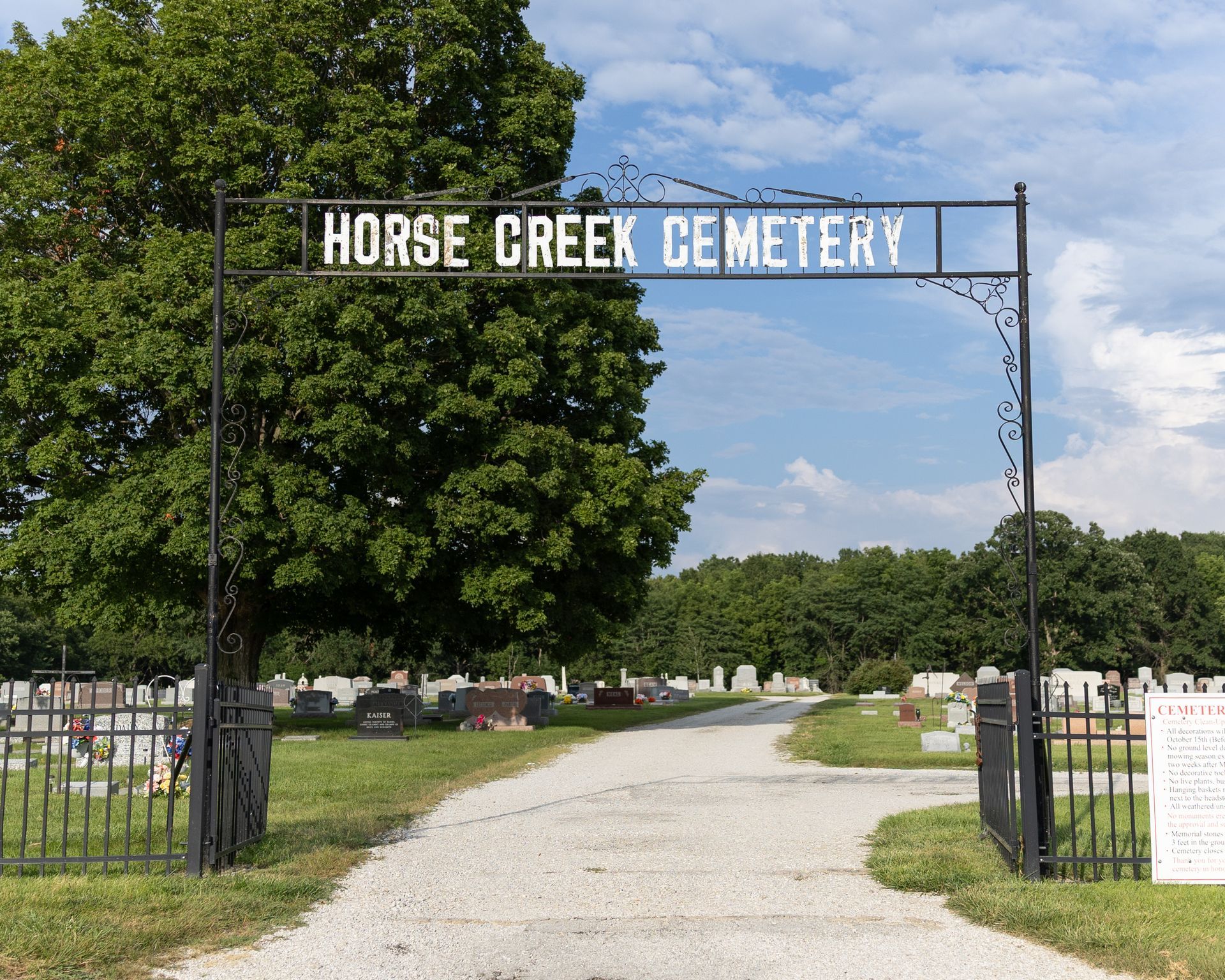 Entrance sign for Horse Creek Cemetery over a gravel path, with trees and gravestones beyond