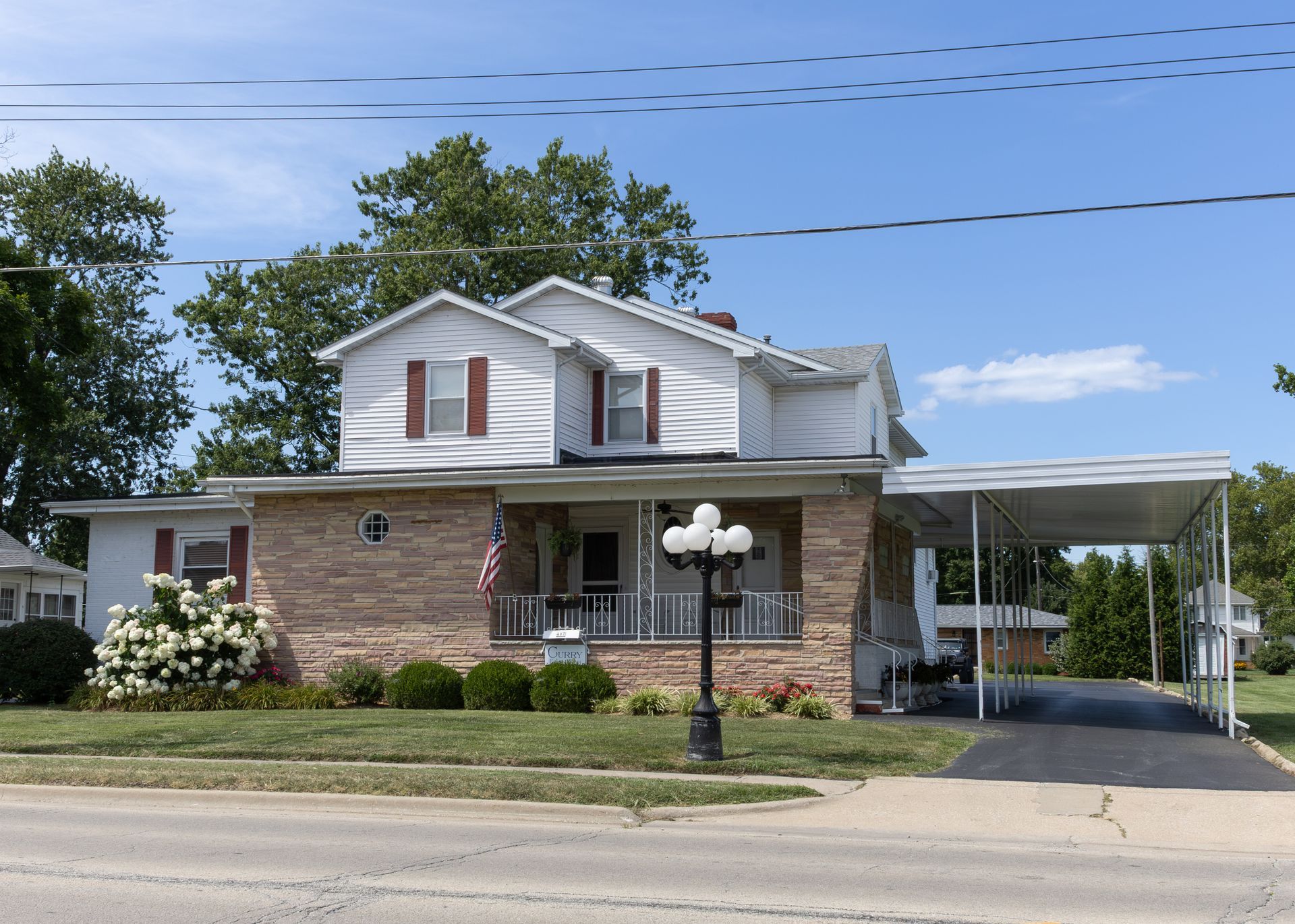 Two-story brick house with white upper siding, front porch, and carport beside a road