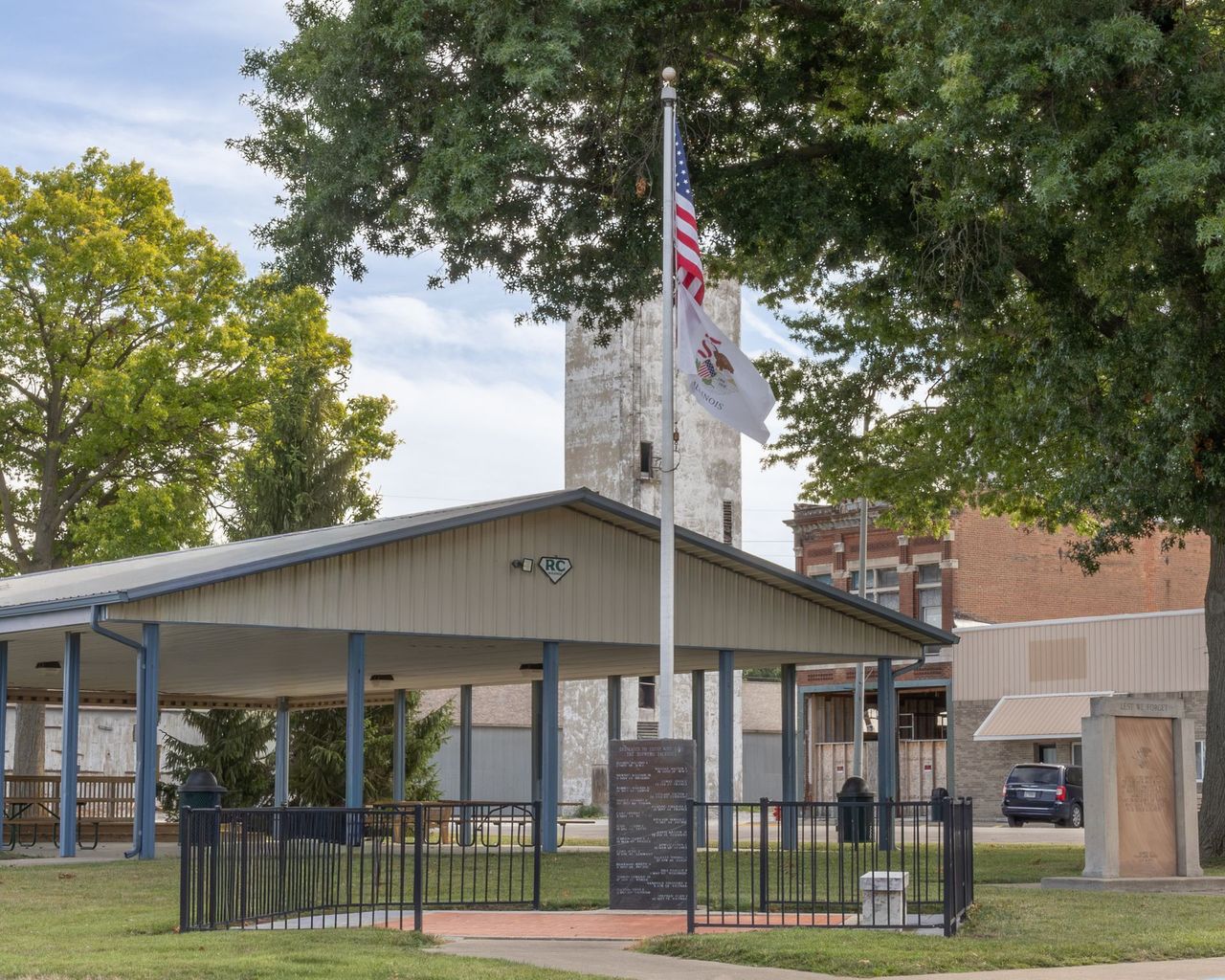 Covered pavilion with flagpoles and fenced memorial in a park-like setting, trees and buildings nearby