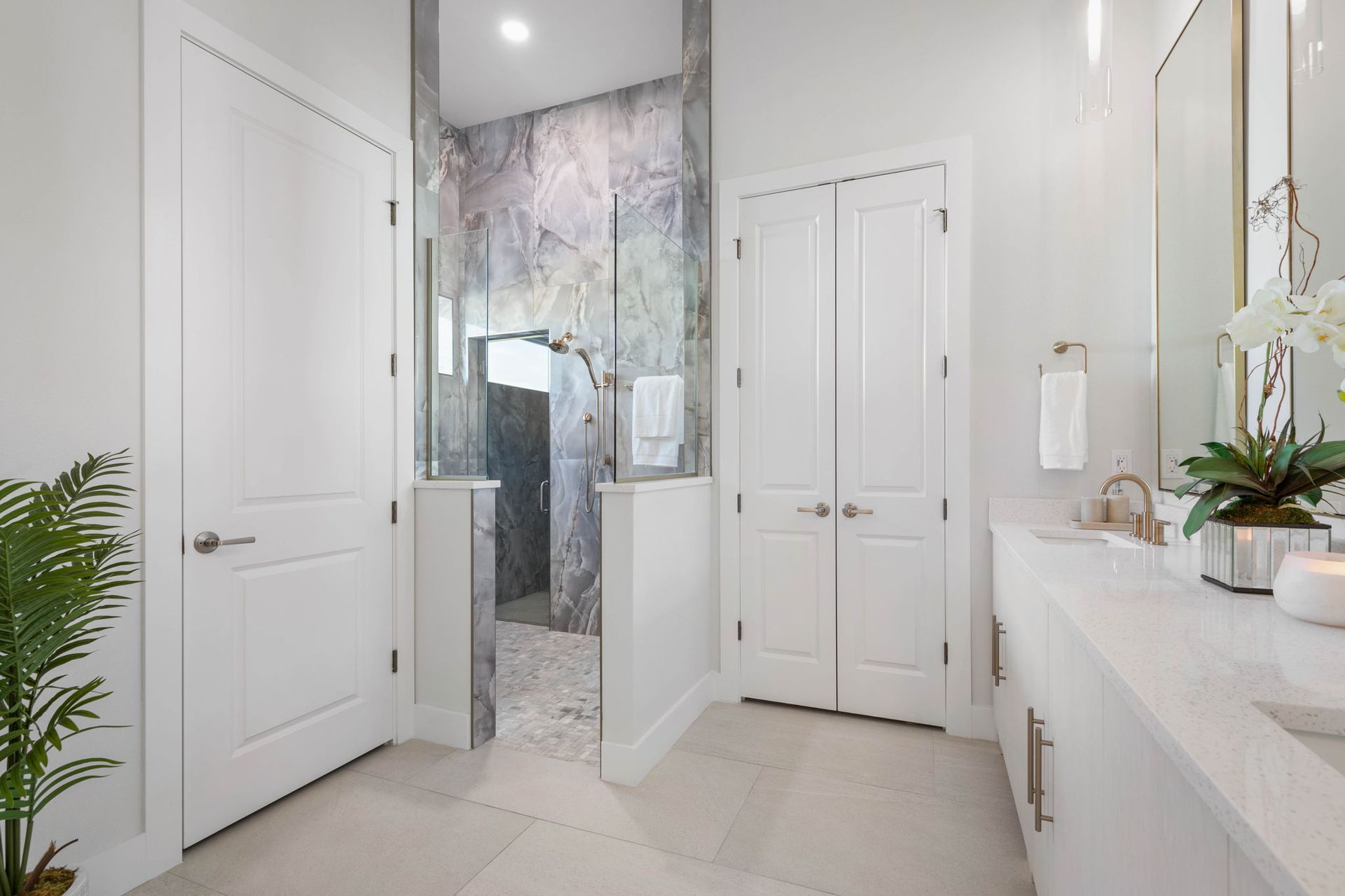 Bright white bathroom with double doors, a glass shower, marble tile, and a vanity with plants.