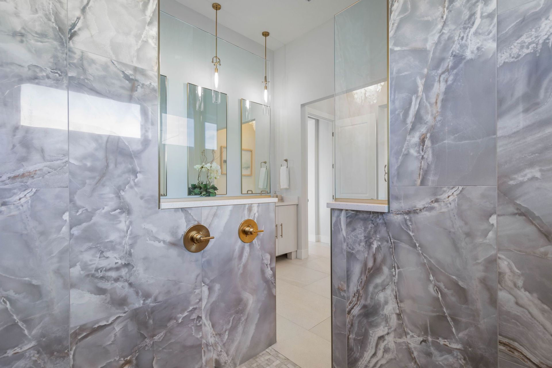 Marble-tiled bathroom shower with glass door and gold fixtures, seen through an open doorway