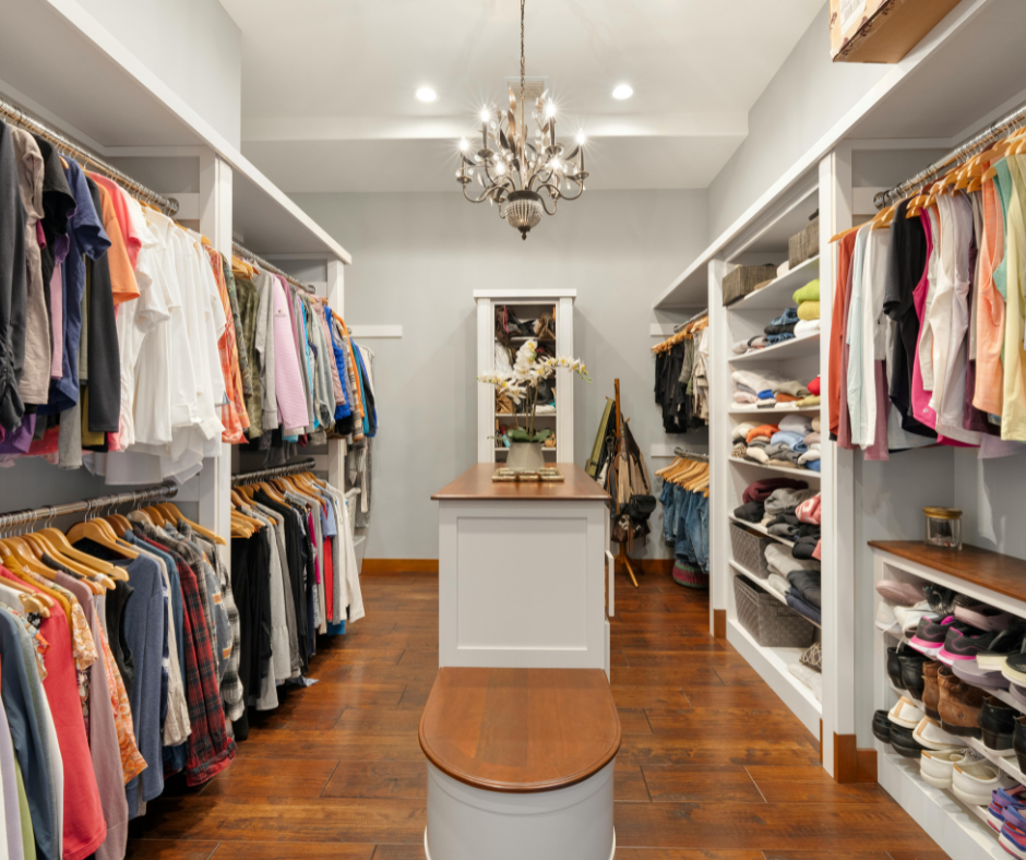 Bright walk-in closet with hanging clothes, shelves, wood floors, and a chandelier.