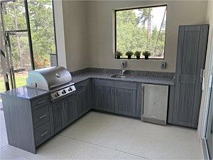 Modern outdoor kitchen with gray cabinets, stainless grill, sink, and large window.