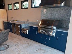 Outdoor kitchen with blue cabinets, stainless steel grill, and stone backsplash under windows