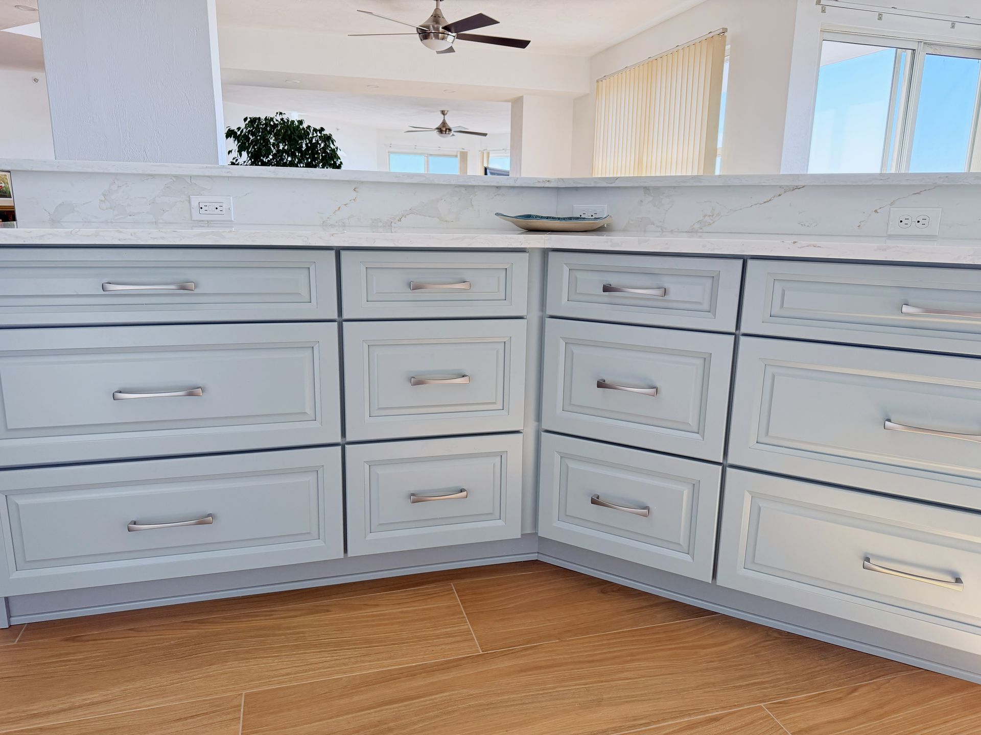Light blue kitchen cabinets with silver handles under a white countertop in a bright room.
