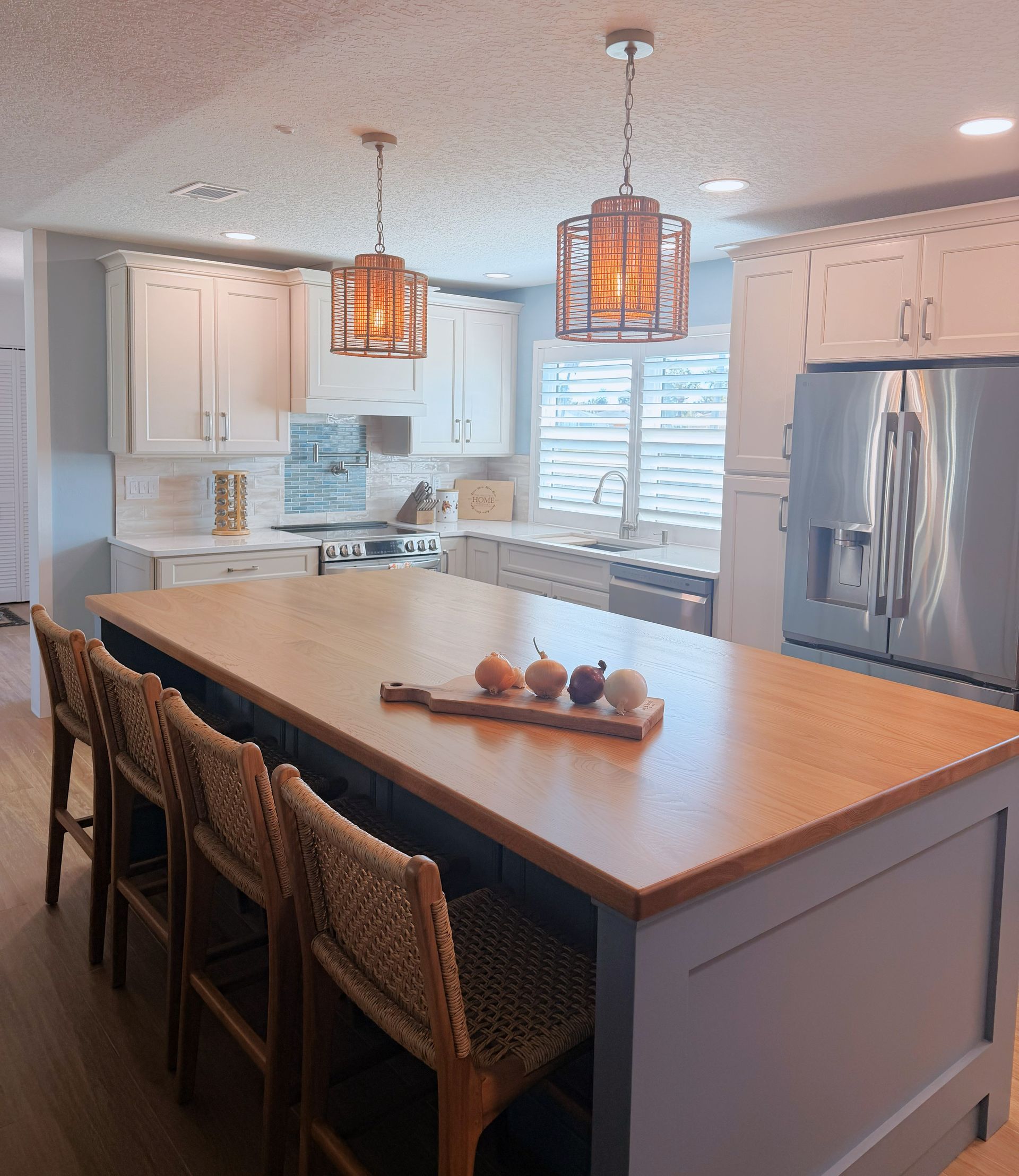 Bright kitchen with large island, woven stools, pendant lights, stainless fridge, and white cabinets.