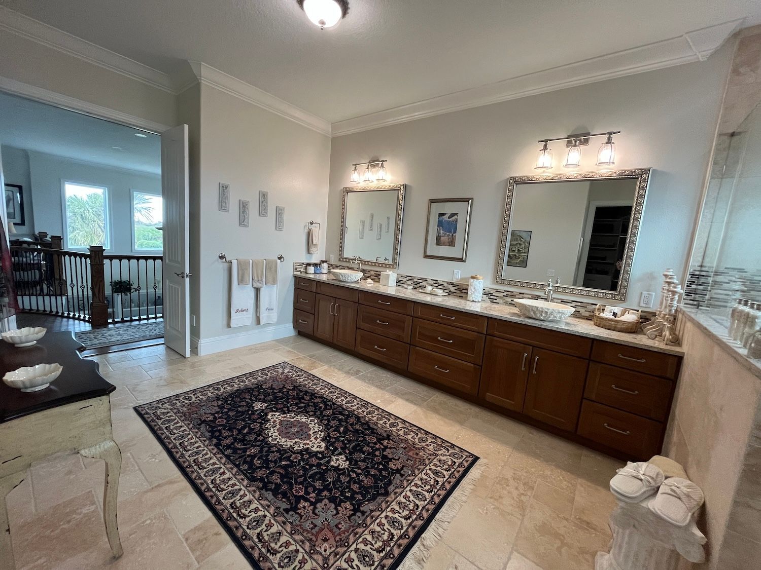 Bright bathroom with double vanity, large mirrors, beige tile floor, and a patterned rug