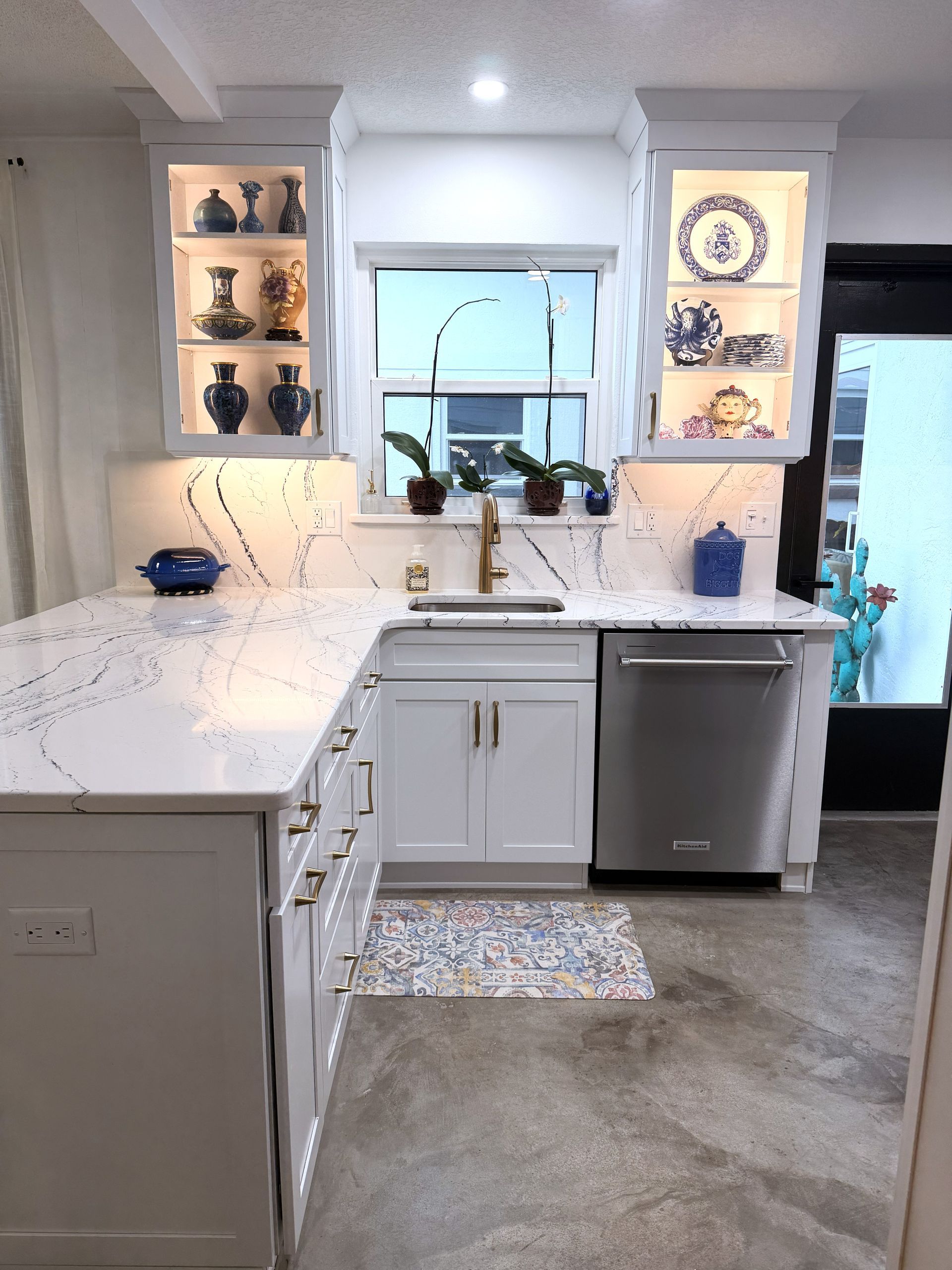 Modern white kitchen with island, sink under window, lit shelves, and stainless dishwasher.