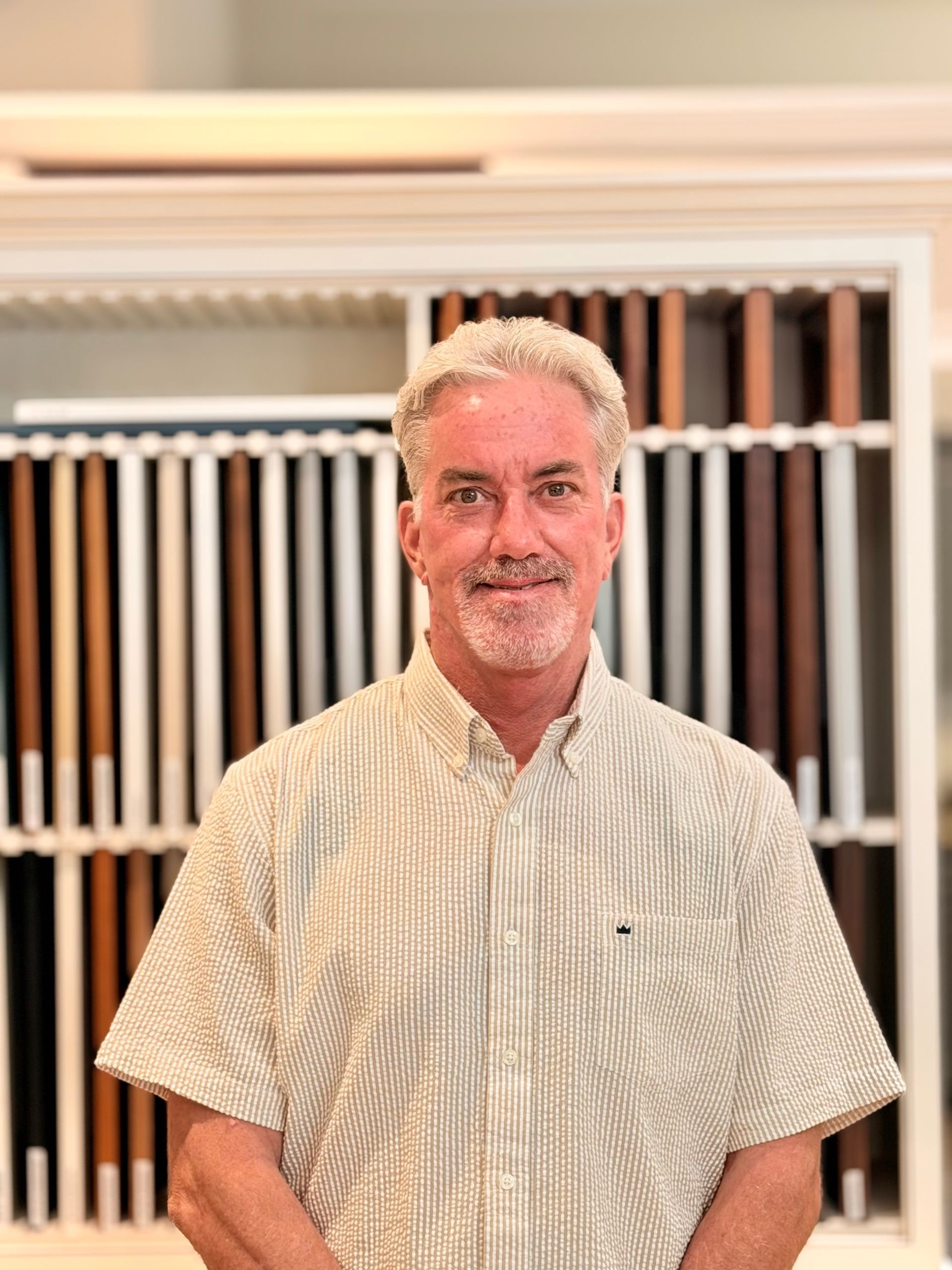 Man standing in a kitchen, smiling in a blue patterned shirt with shelves and jars behind him.