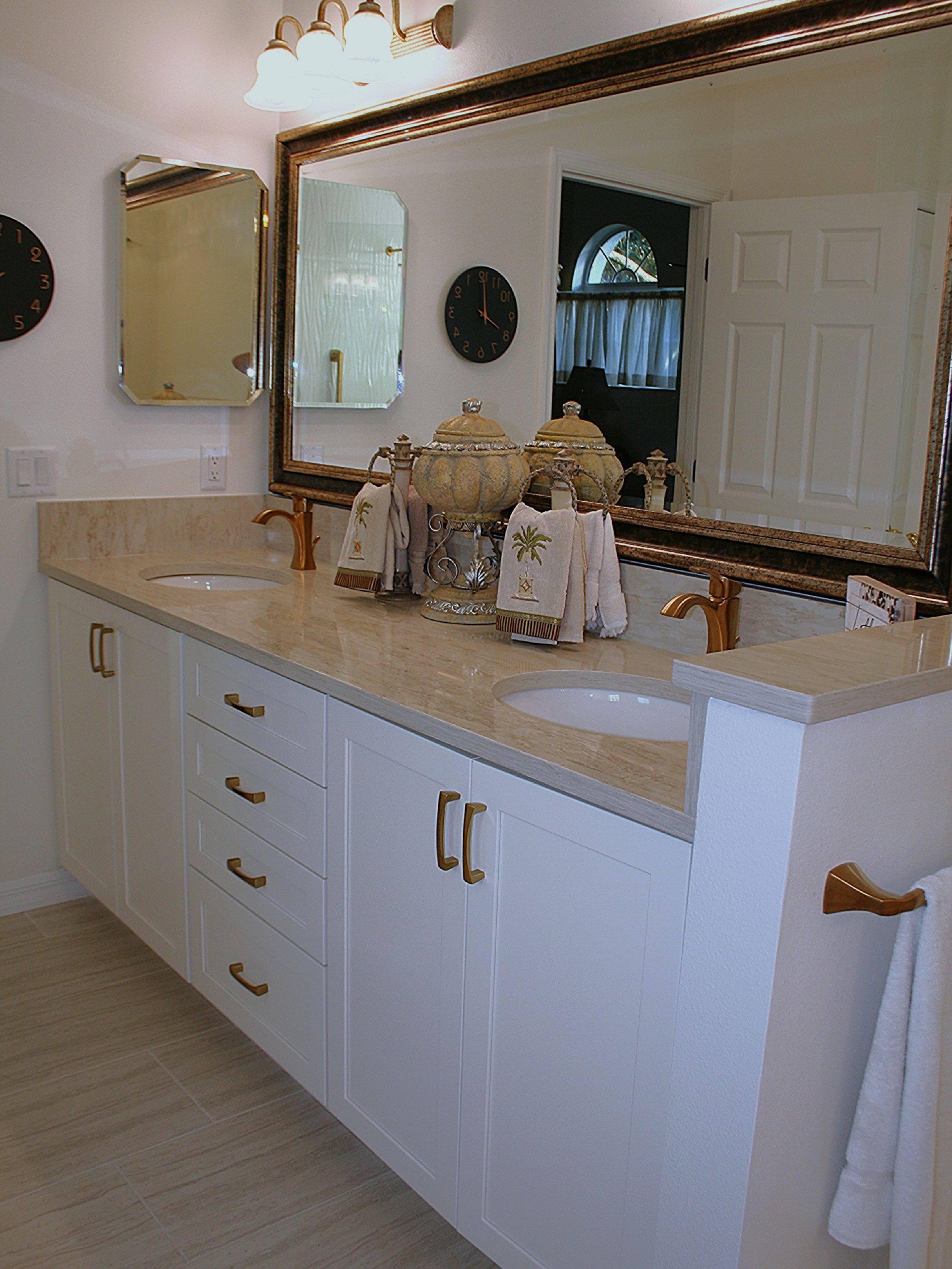 White bathroom vanity with beige countertop, large mirror, and decorative items under a wall sconce.