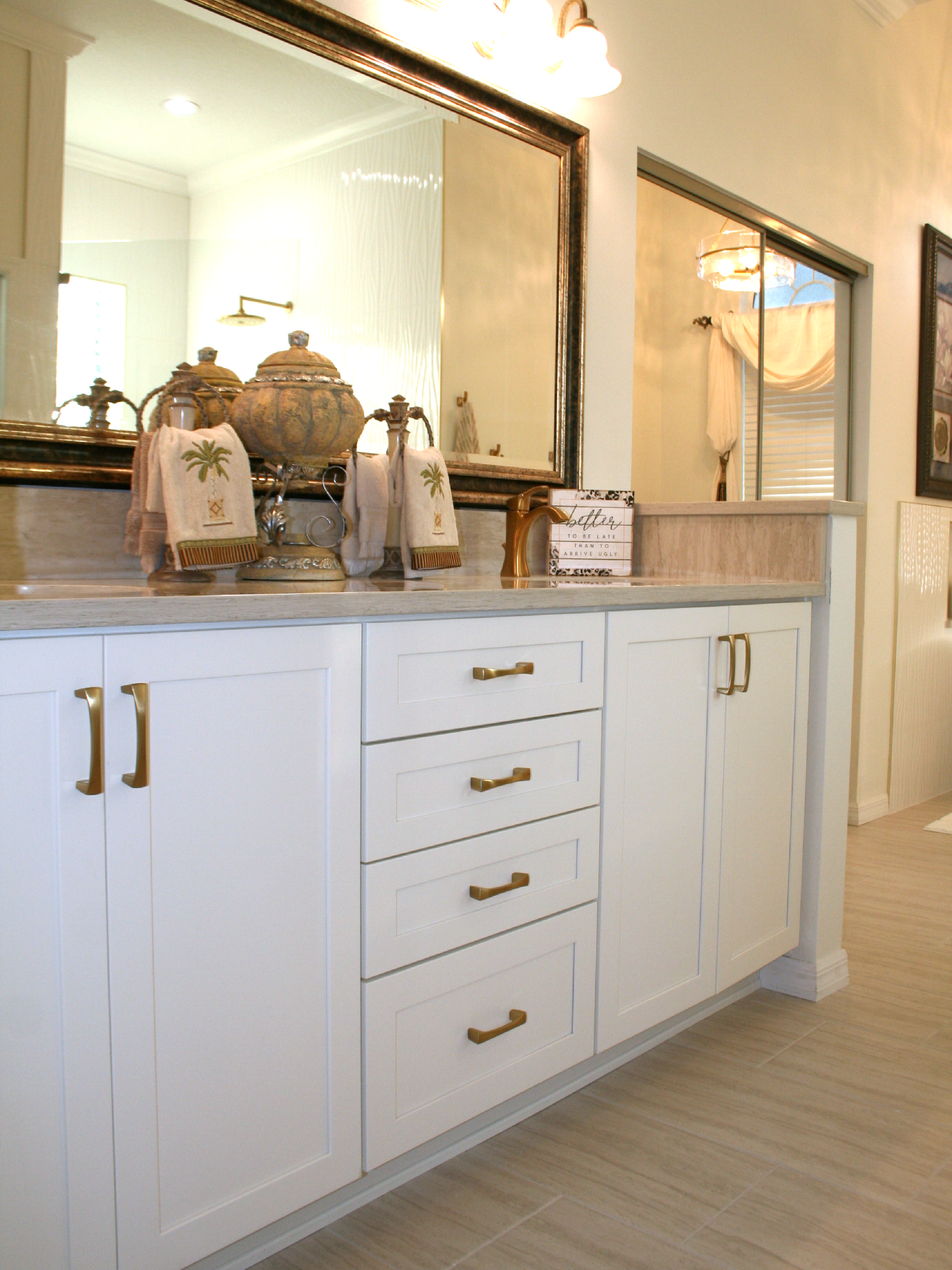 White bathroom vanity with mirror, brass handles, and decorative jars in a bright hallway-style bath