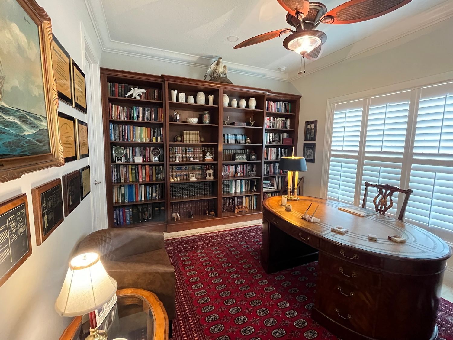 Warm home office with bookshelves, wood desk, red rug, ceiling fan, and paintings on the walls