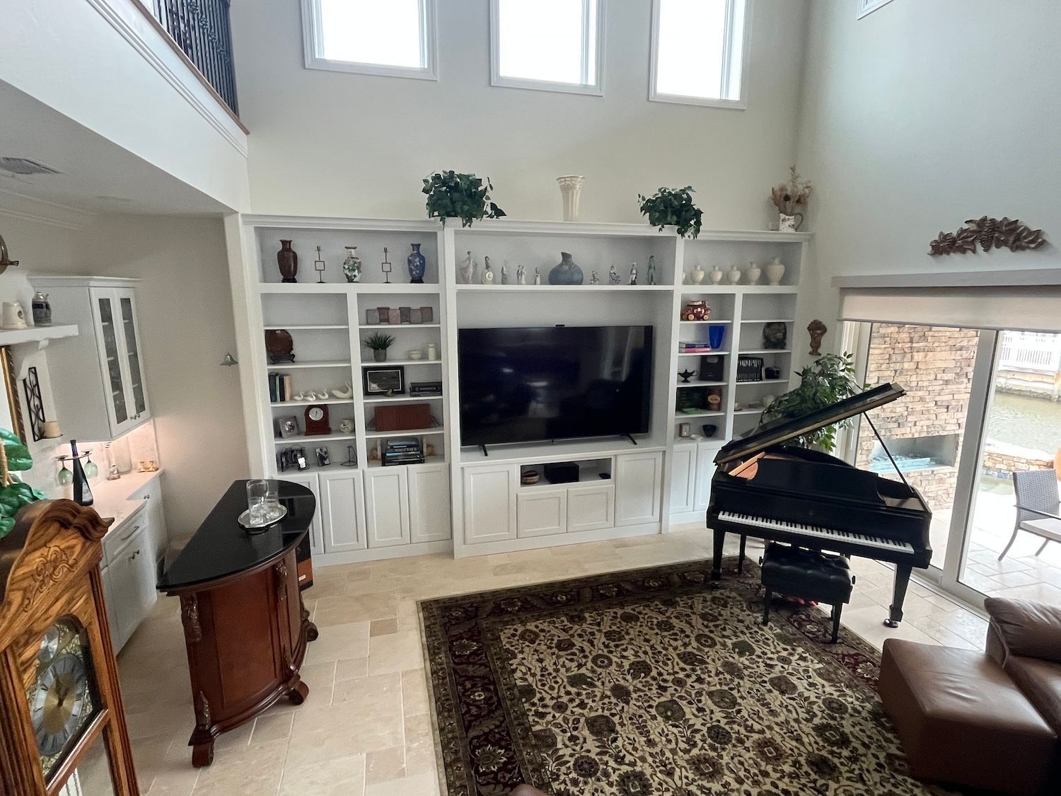 Bright living room with white built-ins, TV, grand piano, and tall windows overlooking trees
