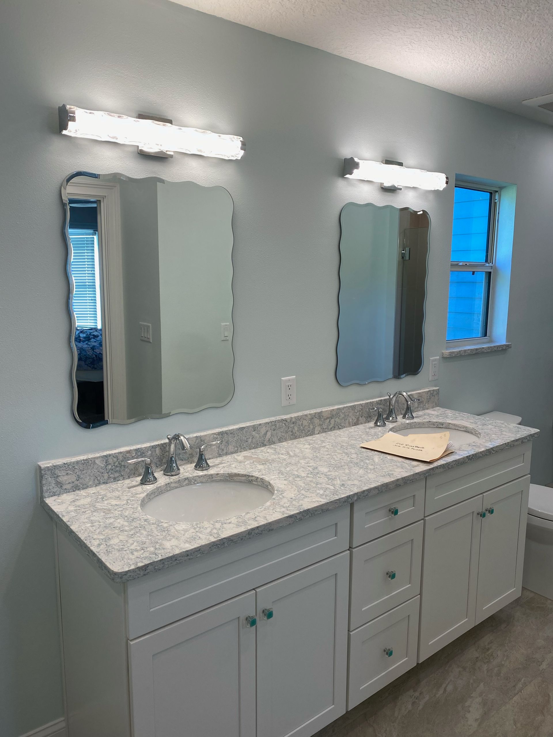 Modern bathroom vanity with two mirrors, bright lights, speckled countertop, and window.