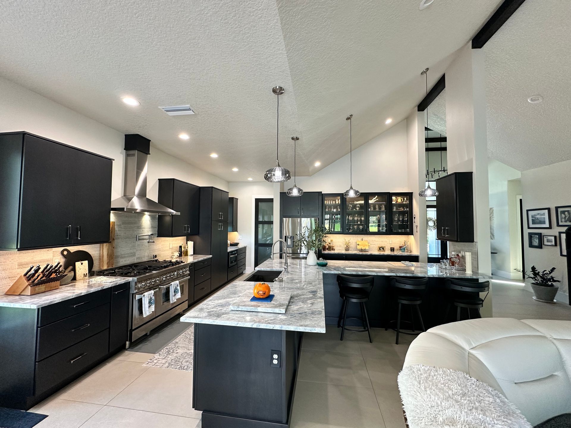 Modern kitchen with dark cabinets, large marble island, pendant lights, and a white leather sofa at right.