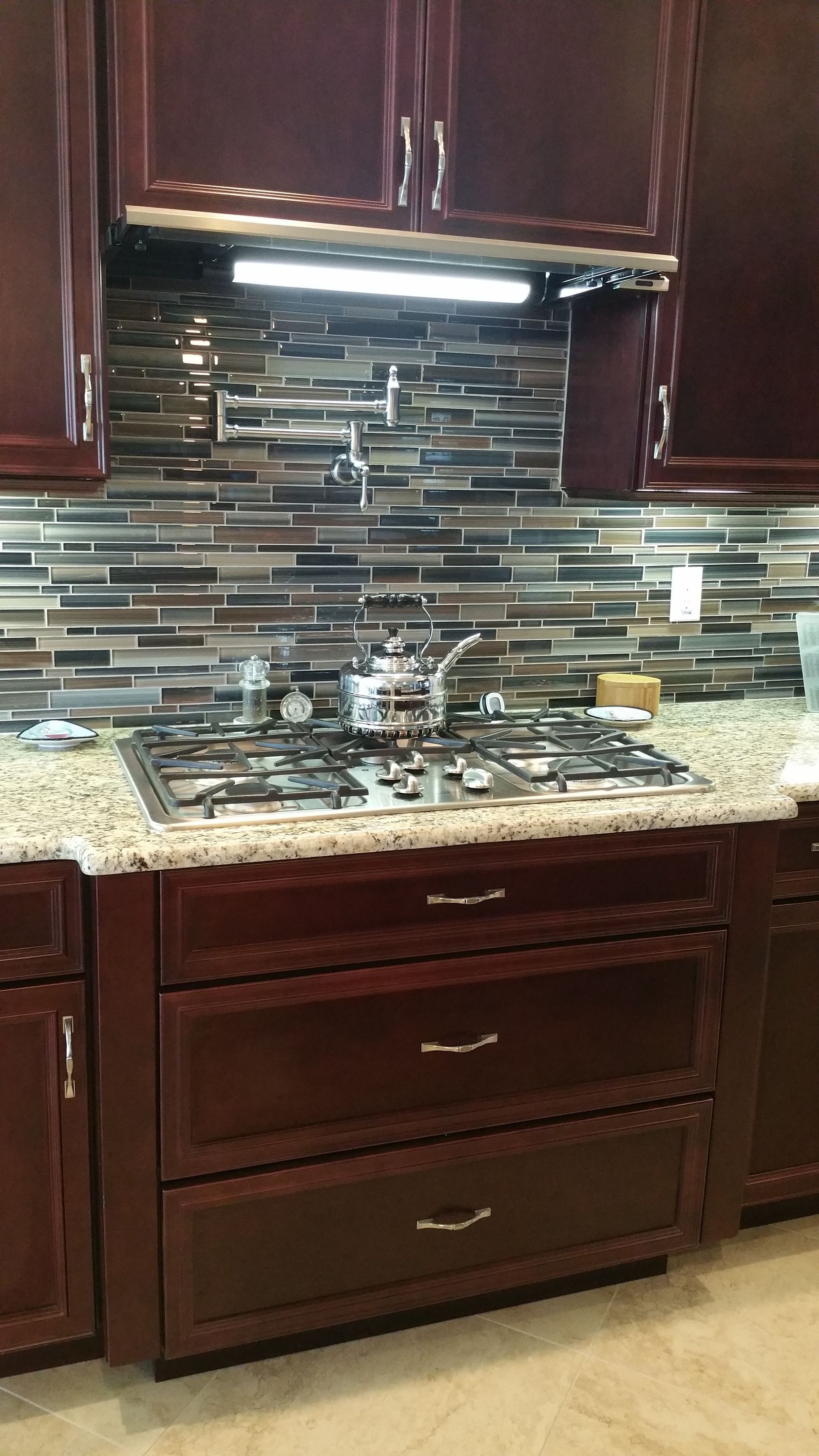 Kitchen stovetop with stainless range hood, mosaic tile backsplash, and dark wood cabinets with granite countertop