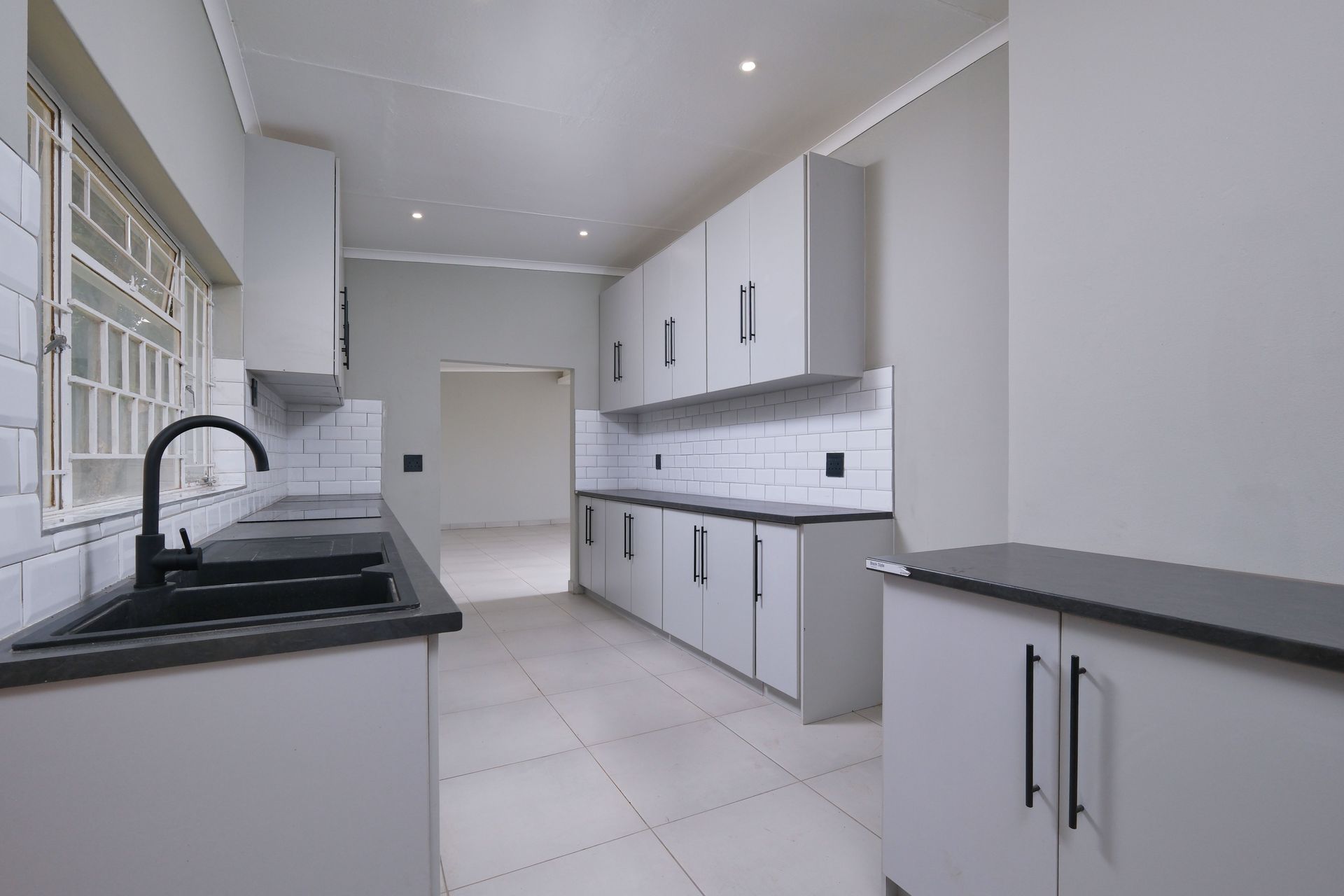 Modern white kitchen with black countertops, tiled floor, cabinets, and large window on the left.