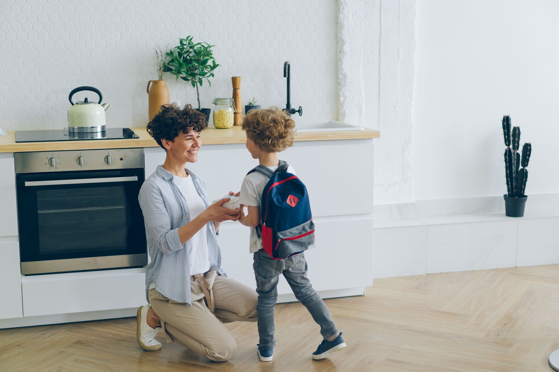 Person kneeling to greet a child in a bright kitchen with white cabinets and a wooden shelf