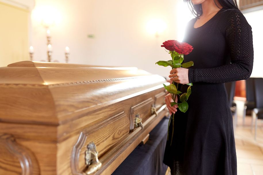 Mourners stand near a floral arrangement. A woman in black weeps, others offer comfort, under umbrellas.
