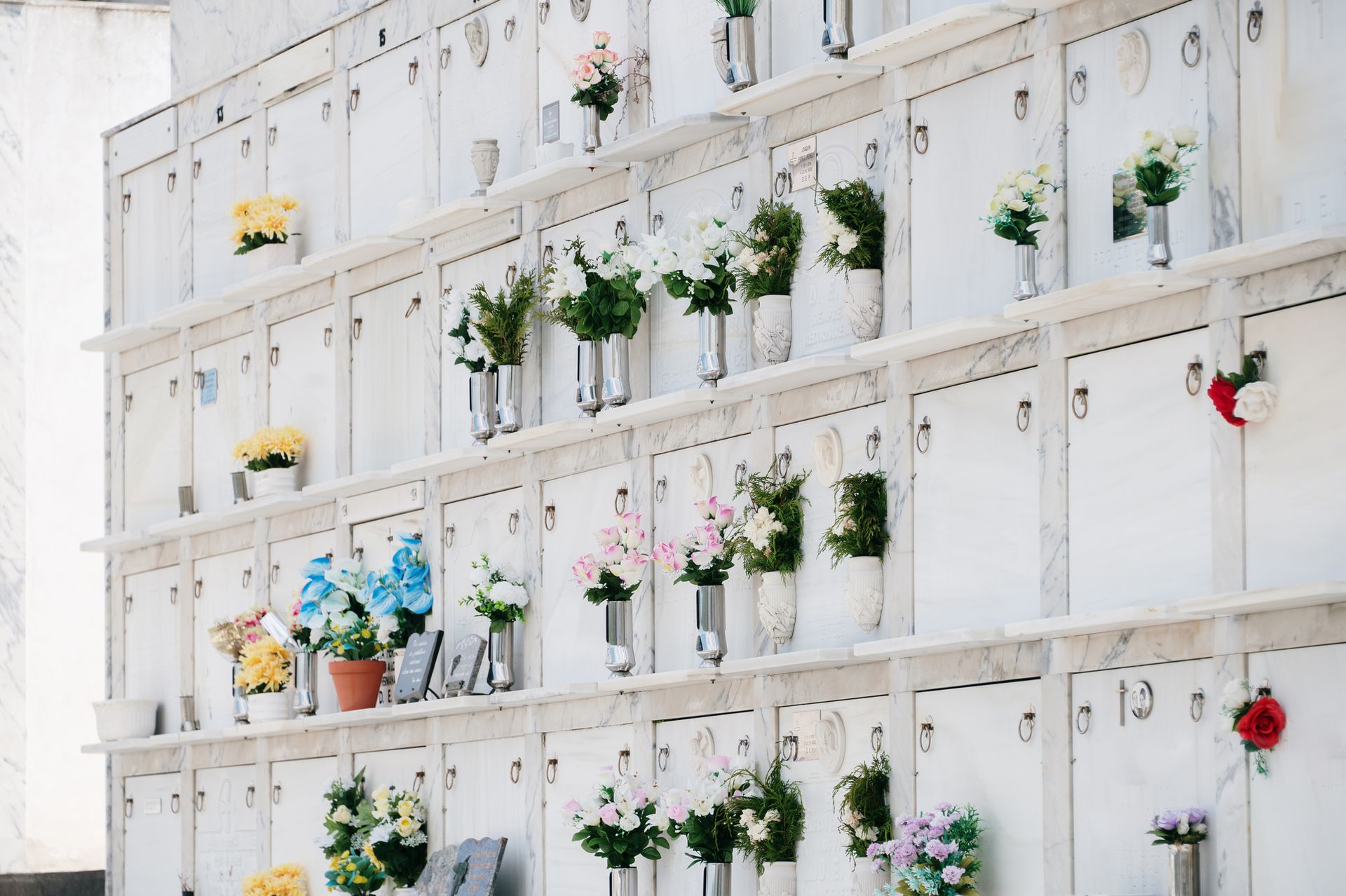 Rows of white columbarium niches, each holding flowers in vases. Rows of white columbarium niches, each holding flowers in vases.