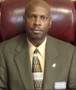 Man in olive suit, beige shirt, patterned tie, seated in a maroon chair, looking forward. Man in olive suit, beige shirt, patterned tie, seated in a maroon chair, looking forward.