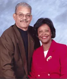 Man in brown blazer, woman in red blazer smile at the camera. Man in brown blazer, woman in red blazer smile at the camera.
