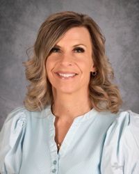 Portrait of a smiling woman with shoulder-length blonde hair wearing a light blue blouse against a gray backdrop