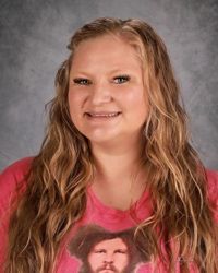 Smiling woman with wavy blonde hair wearing a pink shirt against a gray studio backdrop