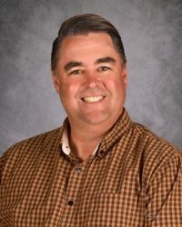 Smiling person in a brown checkered shirt against a gray studio backdrop