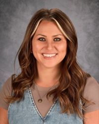 Smiling woman with brown hair in a denim dress against a gray backdrop