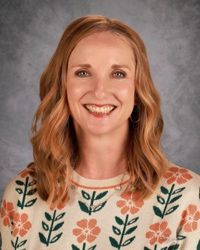 Smiling person with shoulder-length hair in a floral sweater against a gray studio backdrop
